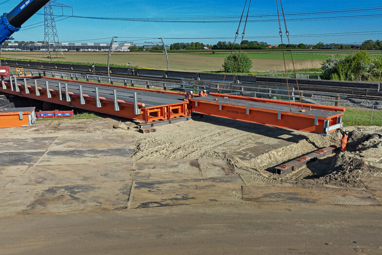 Een oranje brugmodule wordt op zijn plaats getild, met een kraan op de achtergrond.