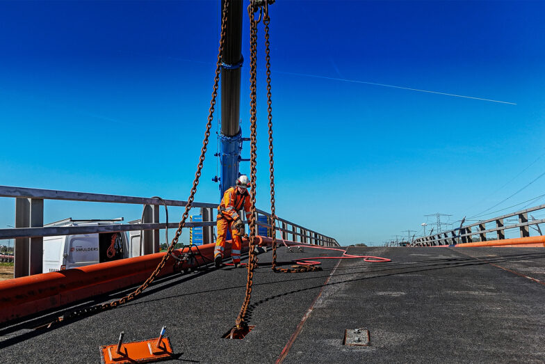 Een oranje retrobridge module wordt op zijn plaats getild door een kraan, op een zonnige dag.
