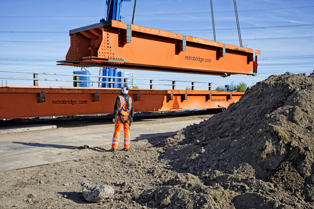 Een kraan tilt een grote oranje retrobridge module op, gezien vanaf een bouwterrein.