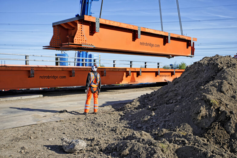 Een kraan tilt een grote oranje retrobridge module op, gezien vanaf een bouwterrein.