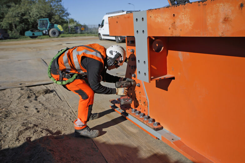 Een arbeider met een veiligheidsharnas en helm maakt een bout vast aan een oranje brugmodule.