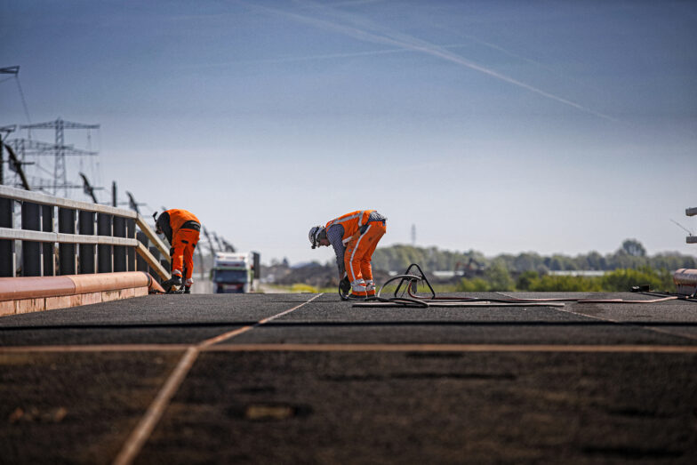 Twee arbeiders in oranje werkkleding leggen een tijdelijk wegdek op een brug.